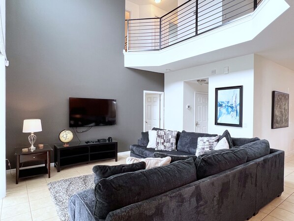 Una sala de estar moderna con un sofá gris, un televisor de pantalla plana, una mesita con una lámpara y una escalera que sube a un nivel superior.