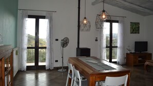 A dining area with a wooden table, white chairs, a fan, and a TV on a wooden cabinet. - Relaxing House surrounded by Olive trees, wonderful sea view (Dolcedo)