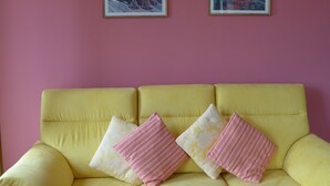 A yellow sofa with three pillows against a pink wall, flanked by two framed pictures.