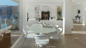 Un salon moderne avec une table à manger en verre entourée de chaises blanches, une cheminée et une vue dégagée grâce à de grandes fenêtres.