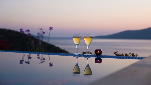 Deux verres de vin blanc posés sur une surface réfléchissante, avec une bougie et des fleurs en arrière-plan.