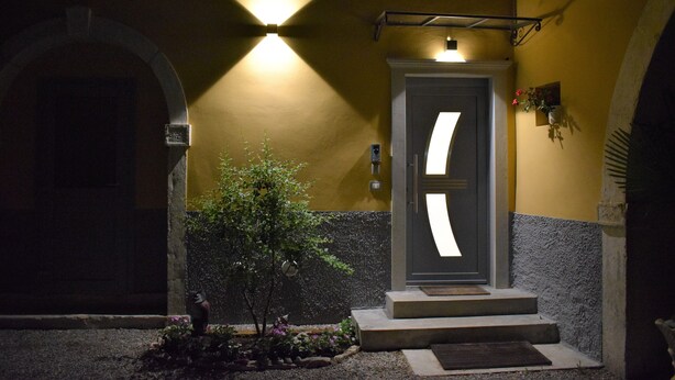 Entrada de un edificio con una puerta iluminada por un foco, un pequeño árbol y un camino de piedra que lleva a la puerta.