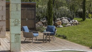 Una terraza de madera con dos sillas de mimbre y una mesa pequeña, una piscina y un edificio de madera con una puerta al fondo.