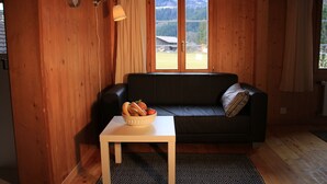 Una habitación con paneles de madera, un sofá de cuero negro, una mesa blanca con un cuenco de pan y fruta, y una ventana que ofrece vistas a una montaña y una casa.