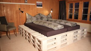 Una habitación con paneles de madera, una cama hecha con palets blancos, una silla y un escritorio.