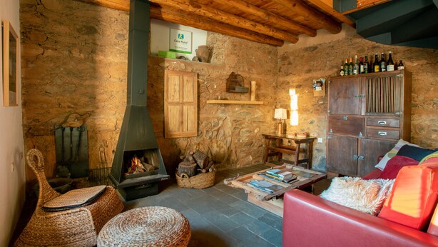 Un interior rústico con pared de piedra, techo de madera, chimenea, un aparador de madera con botellas y un sofá rojo.