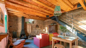 Un interior rústico con vigas de madera a la vista, pared de piedra, chimenea, sofá rojo y zona de comedor con taburetes.