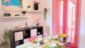 A dining area with a table set for two, a kitchen counter, a potted plant, and a window with pink curtains. - Apartment Salgari Ponente (Marina di Ragusa)