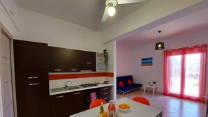A modern kitchen with dark cabinets, a white countertop, and a dining area with a table set for two. - Apartment Salgari Ponente (Marina di Ragusa)