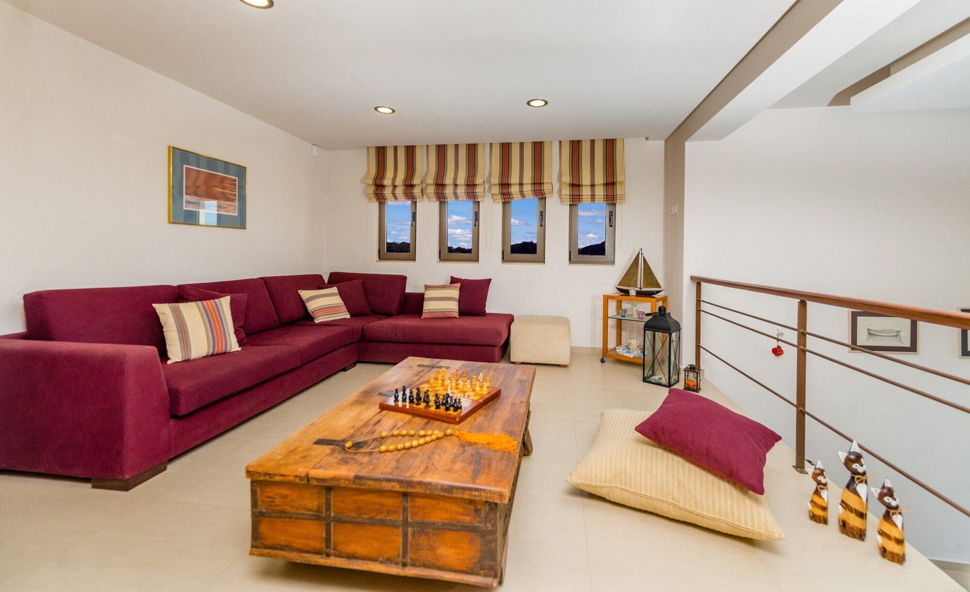 A living room with a sectional sofa, a wooden coffee table with chess pieces, a small ottoman, and a view of the outdoors through windows.