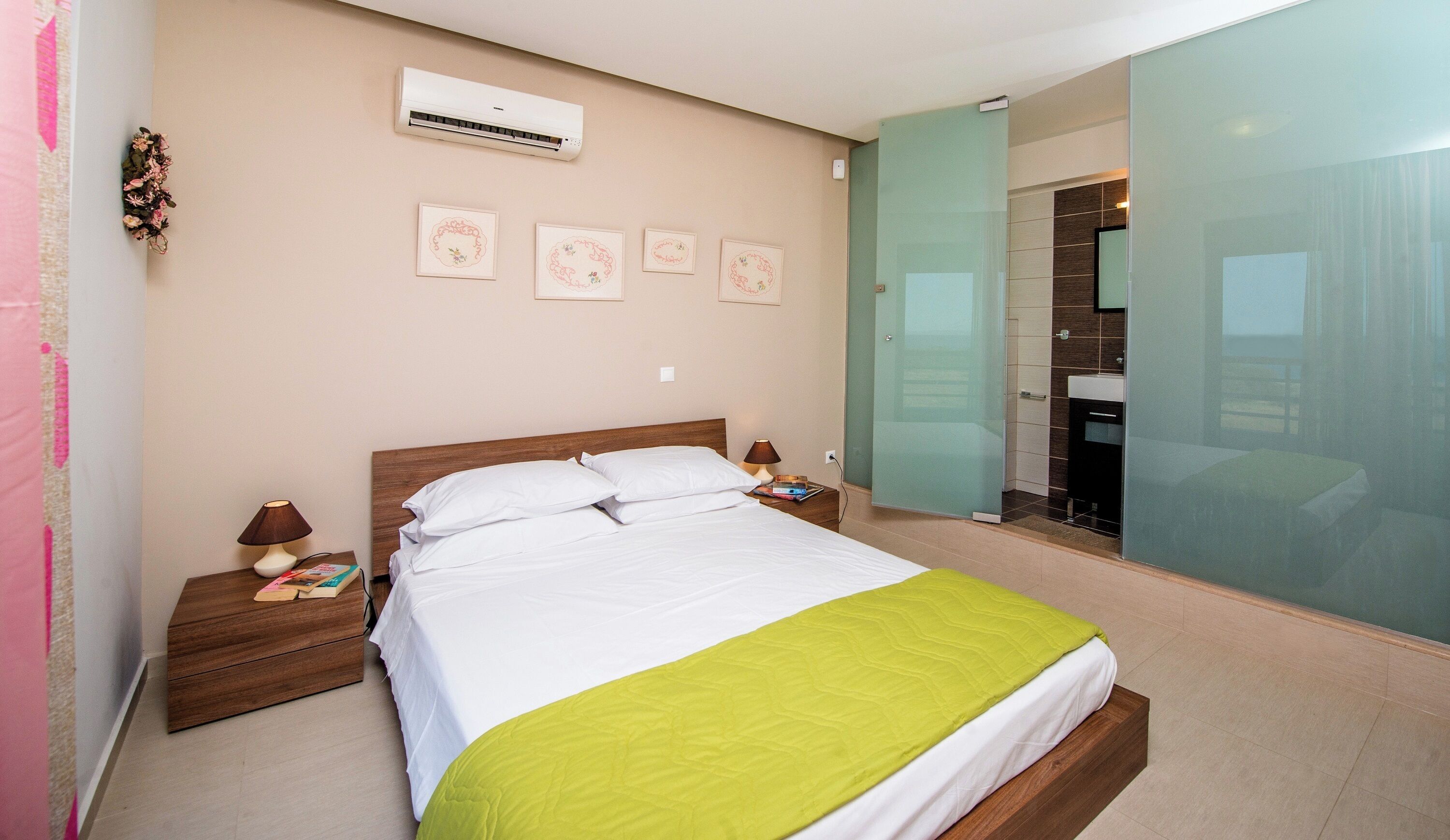 A bedroom with a bed, bedside tables, an air conditioner, and a bathroom with a sink and mirror.