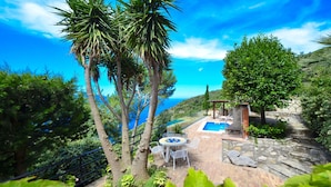 A pool area with a stone patio, white metal chairs, and a view of the sea.