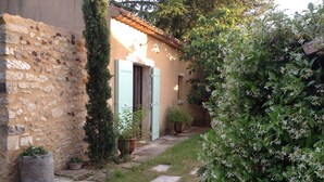 A narrow stone pathway leading to a rustic building with a tiled roof, a wooden door, and a small garden area with potted plants and flowering shrubs. - Les Mûriers: 55m2 gite for 2/4 people in a quiet location overlooking the Luberon and vineyards (Gargas)