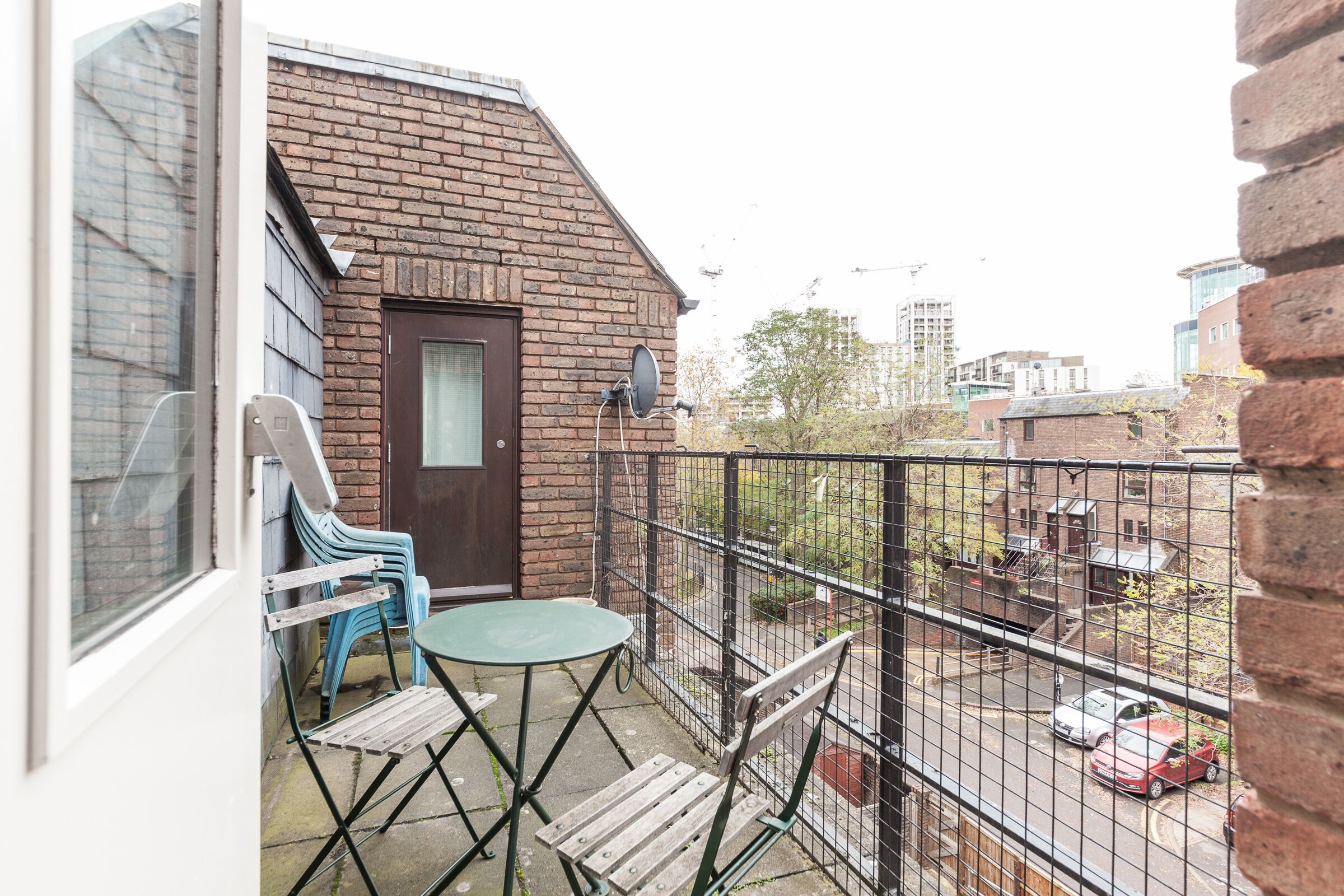 A balcony with a brick wall, a door, and a metal fence. There are two chairs and a table.