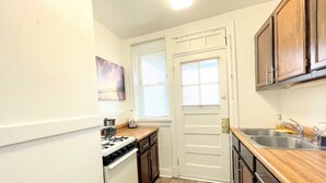 A compact kitchen with white cabinets, a stove, a sink, and a window with blinds. - 6649 2W | Central St. Louis Beauty (St. Louis)