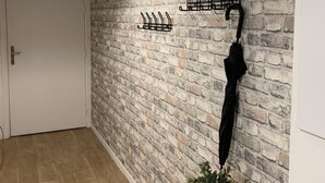Um corredor com parede de tijolos, luminária pendente, suporte para casacos, um guarda-chuva e uma pequena mesa com uma planta.