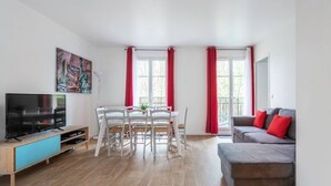 Uma sala de estar moderna com uma TV de tela plana, uma mesa de jantar com cadeiras, um sofá cinza com almofadas vermelhas e janelas grandes com cortinas vermelhas.