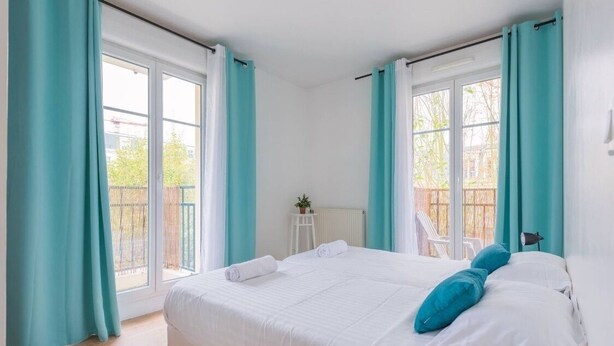 Quarto com uma cama grande, cortinas verde-água, uma mesinha com uma planta e vista para a varanda.