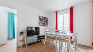 Uma sala de estar moderna com uma televisão de tela plana, uma mesa de jantar com cadeiras brancas, uma cortina vermelha e um quadro da Estátua da Liberdade.
