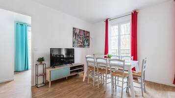 Un salon moderne avec un téléviseur à écran plat, une table à manger avec des chaises blanches, un rideau rouge et un tableau représentant la Statue de la Liberté.