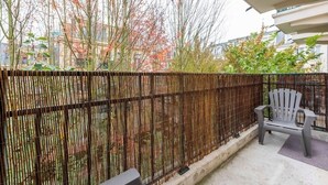 Uma varanda com cerca de bambu, uma cadeira de plástico cinzenta e vista para prédios e árvores.