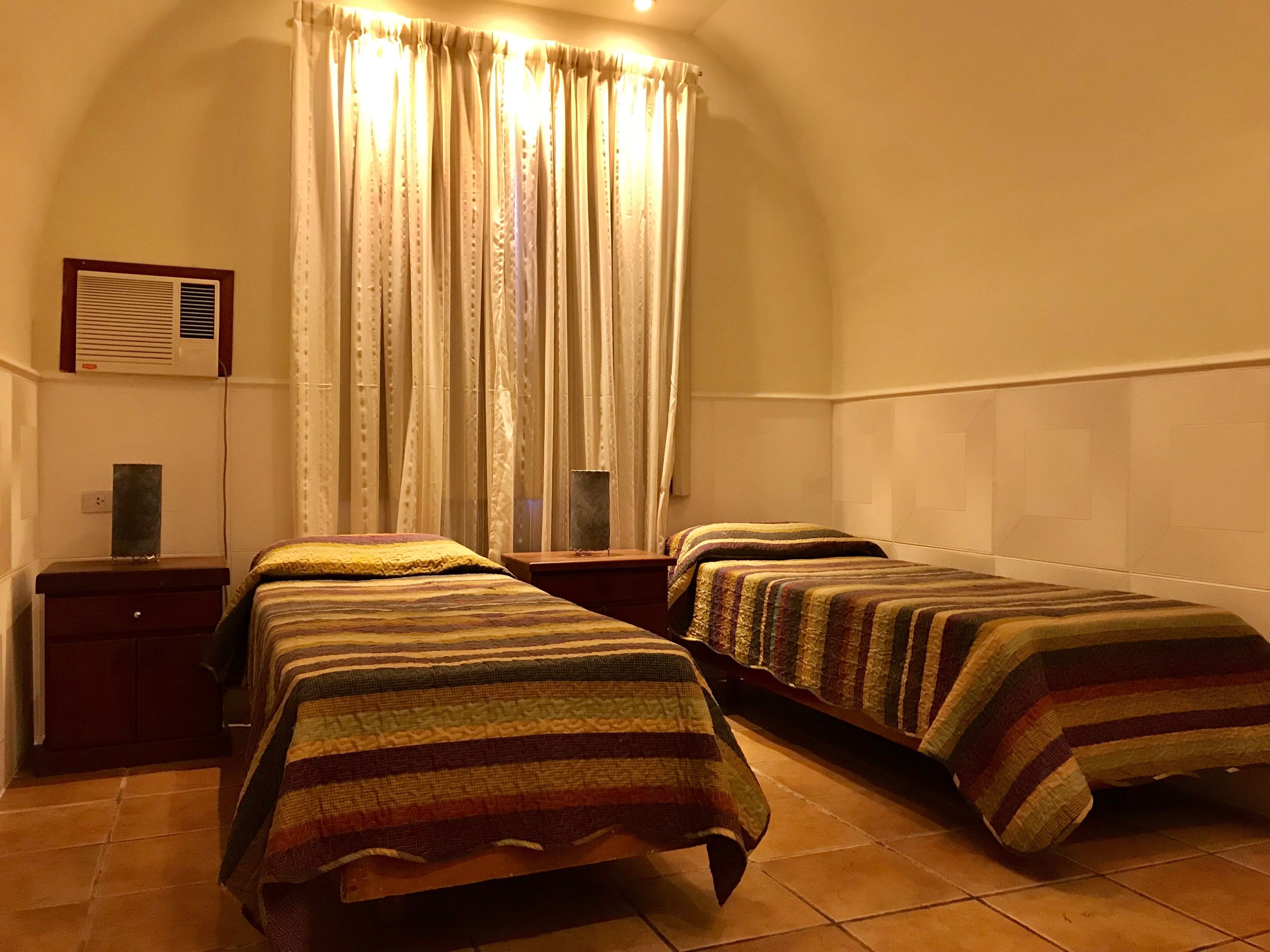 Una habitación con dos camas, una mesita pequeña, aire acondicionado de pared y un ventanal con cortinas.