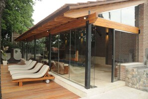A modern outdoor lounge area with a wooden pergola, glass walls, and a stone fireplace. - Hotel Riviera (Villa Carlos Paz)