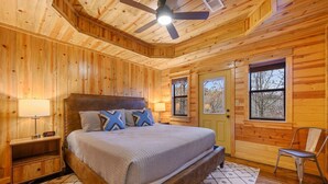 Un dormitorio de cabaña de madera con cama, mesitas de noche, una silla y un ventilador de techo.