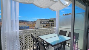 Un balcón con una mesa y sillas de vidrio, celosía blanca y vistas a edificios y colinas.