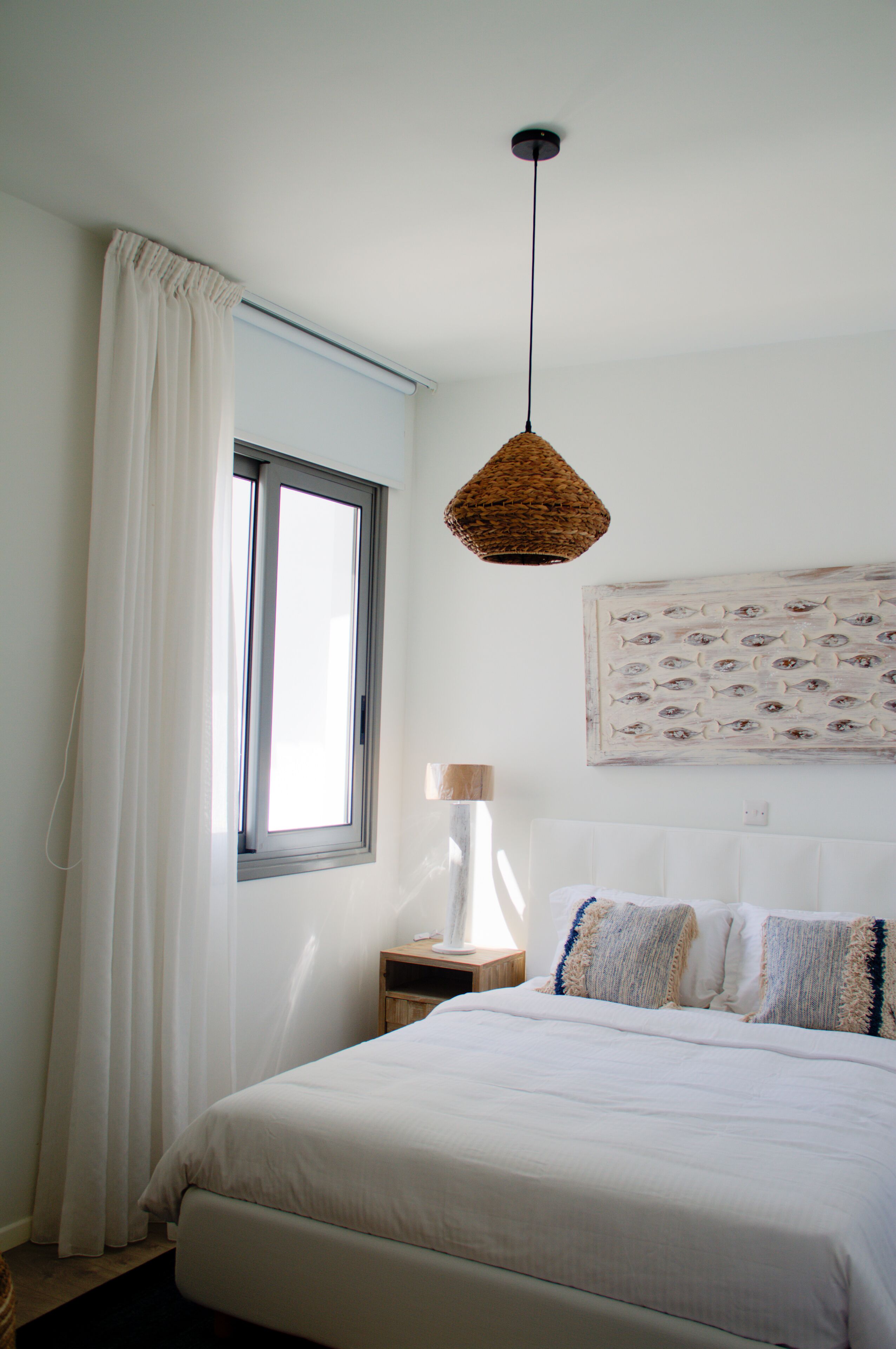 A bedroom with a bed, a bedside table, a lamp, a hanging light fixture, a window with curtains, and a wall art piece.