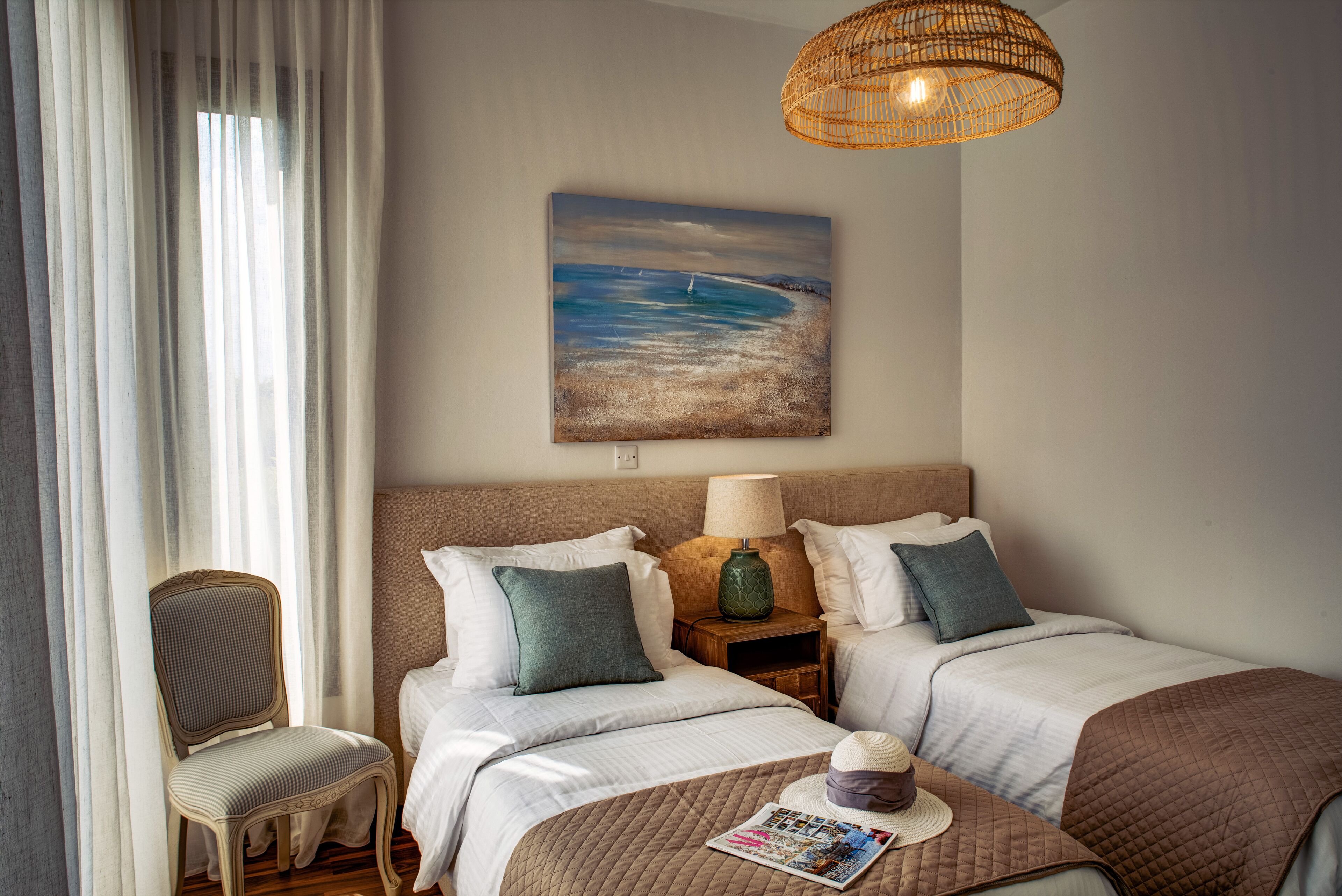 A hotel room with two beds, a chair, a lamp, and a painting of a beach scene.