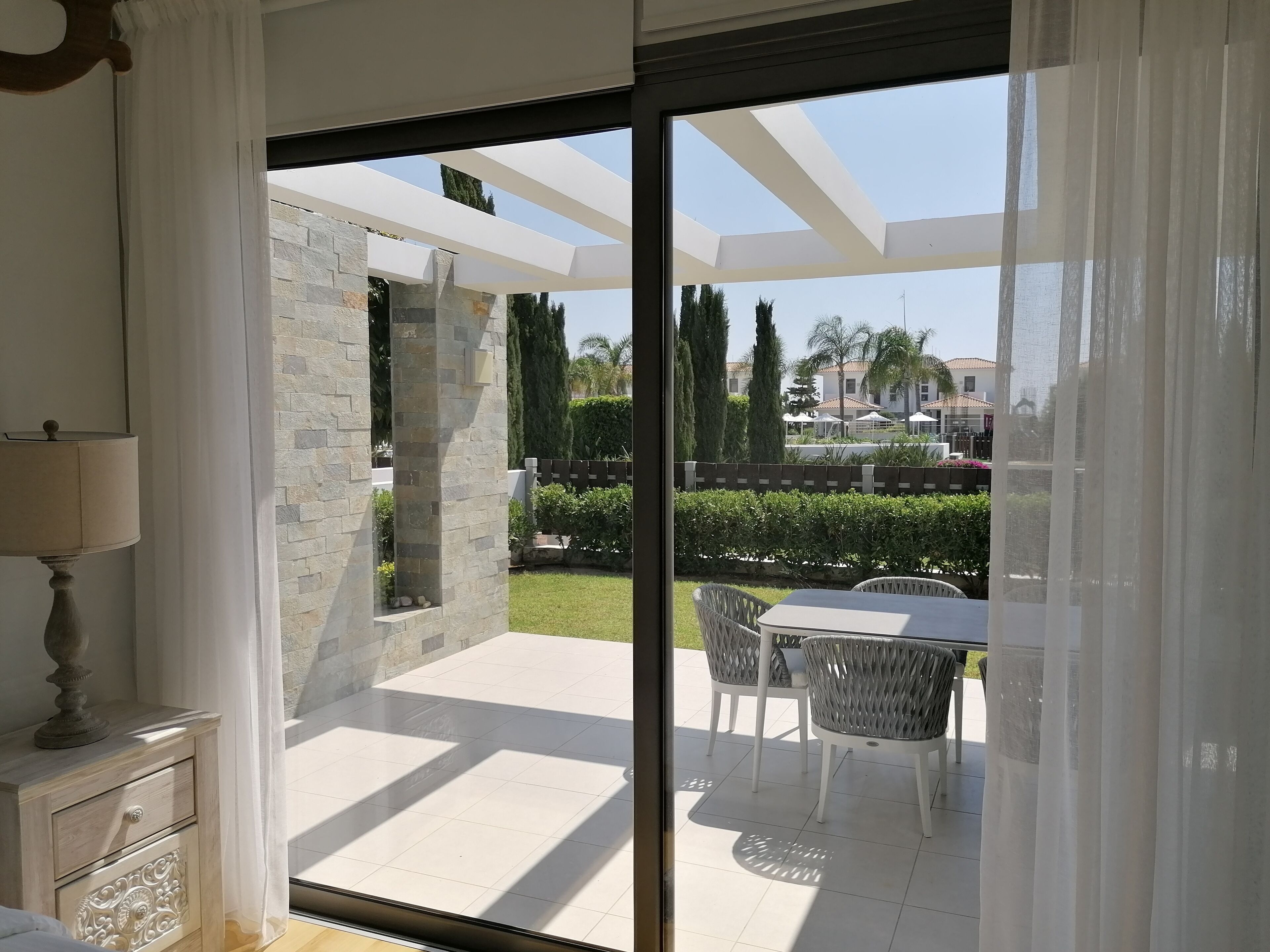 A modern room with a sliding glass door leading to an outdoor patio. The patio features a stone pillar, a white table with wicker chairs, and a view of a well-maintained garden and neighboring buildings.