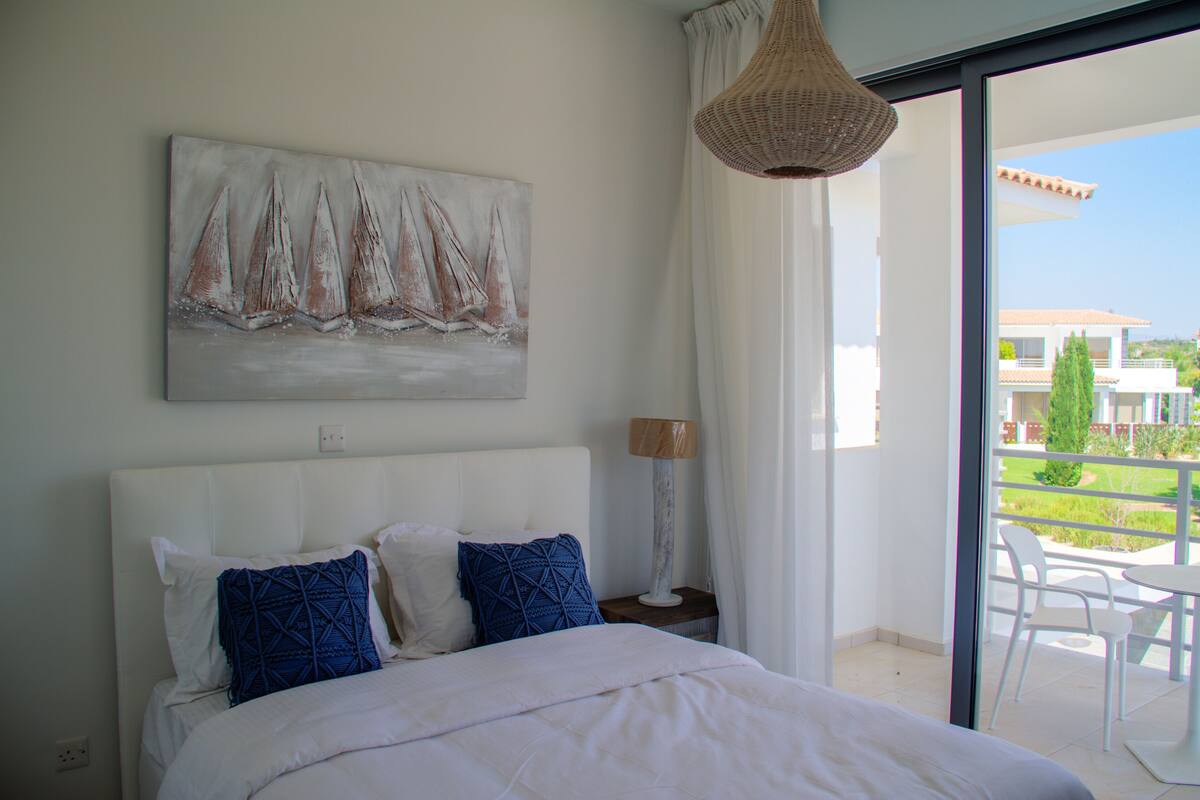 Ein Schlafzimmer mit einem großen Bett, einem Gemälde von Segelbooten, einem Nachttisch mit Lampe und Blick auf einen Balkon mit Gartenmöbeln.