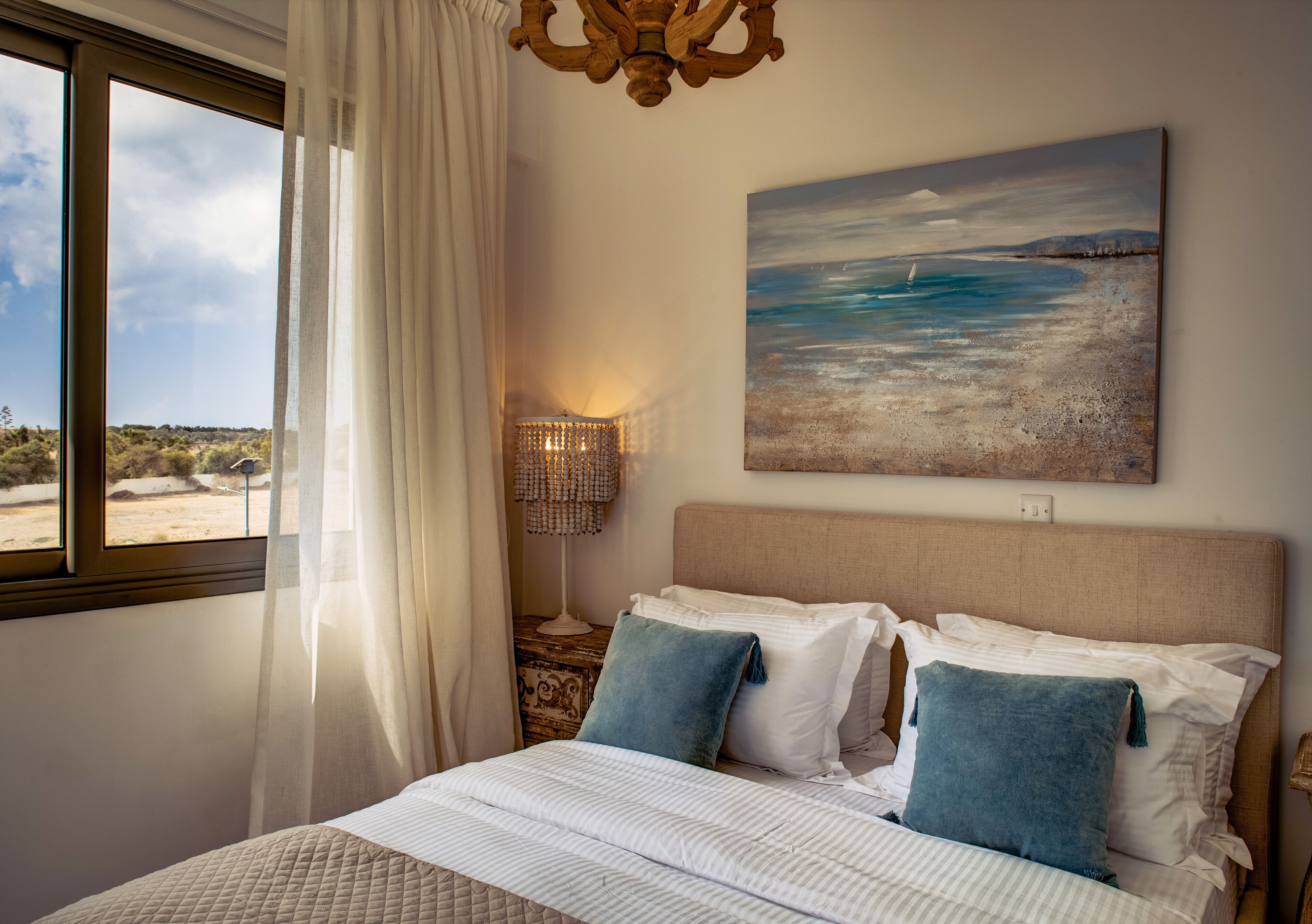 Una camera da letto con un letto grande, un quadro raffigurante una spiaggia, una lampada da comodino e vista sull'esterno attraverso una finestra.
