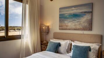 A bedroom with a large bed, a painting of a beach scene, a bedside lamp, and a view of the outdoors through a window.