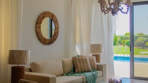 A modern living room with a sofa, coffee table, and dining table. There is a large mirror on the wall and a view of a swimming pool through the glass door. - Atlantis Gardens (Mazotos)