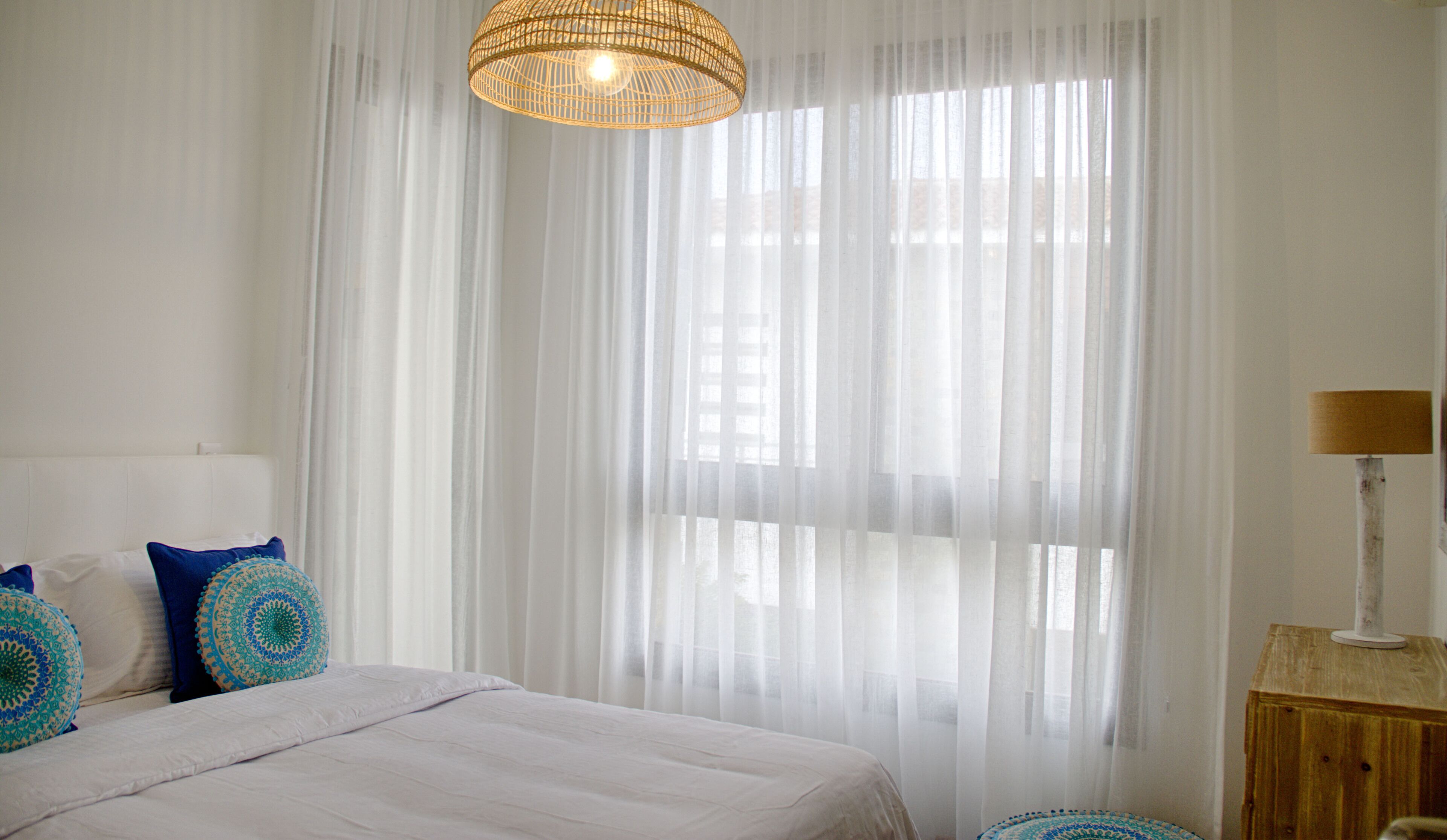 Un dormitorio con cama, cortinas blancas, lámpara colgante y mesita de noche con lámpara.