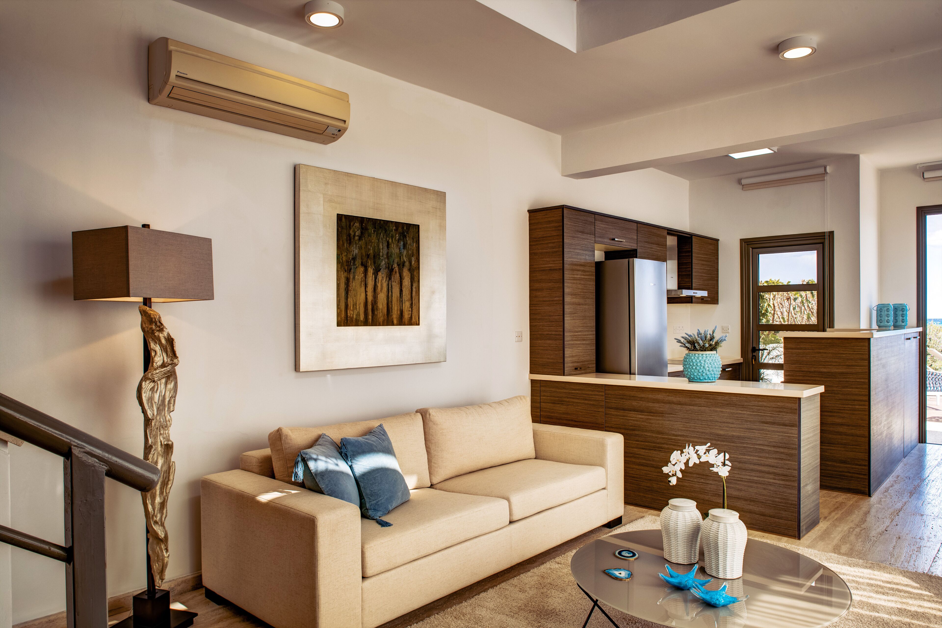 A modern living room with a beige sofa, a glass coffee table, and a kitchen area with wooden cabinets and a refrigerator.