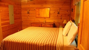 A wooden cabin room with a bed, a nightstand, a lamp, and a window with curtains.