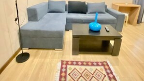 A modern living room with a grey sectional sofa, a black coffee table, a colorful rug, a floor lamp, a painting on the wall, and an air conditioning unit. - Downtown Hotel Tbilisi (Tbilisi)