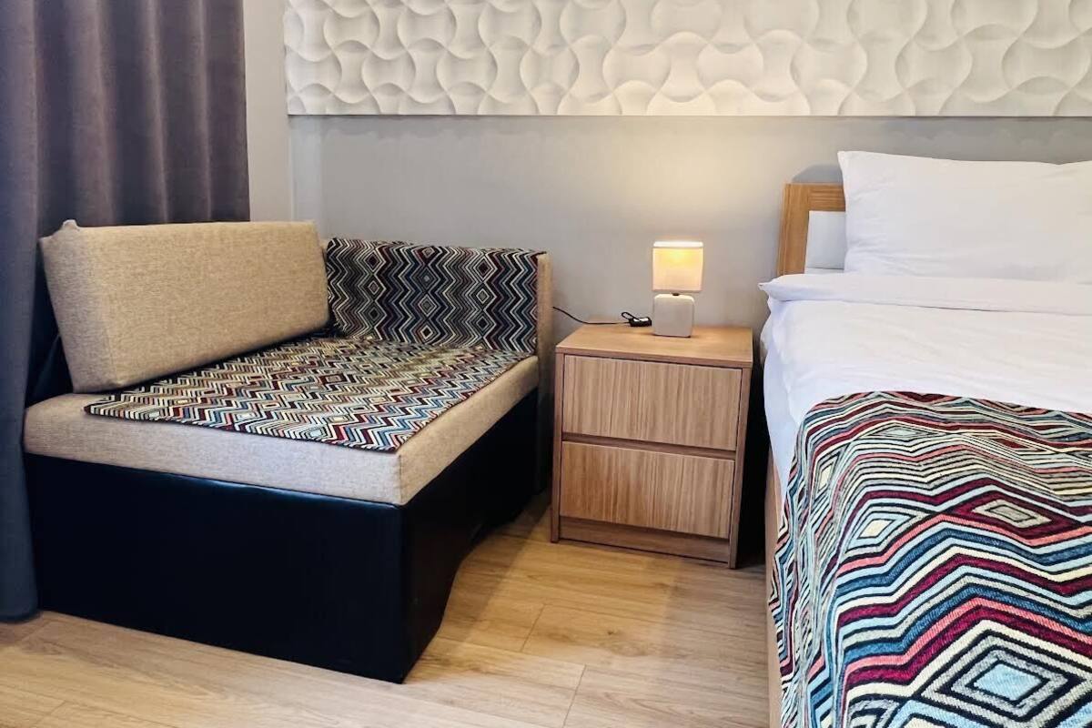 A hotel room with a sofa, a bedside table with a lamp, and a bed with a patterned blanket.