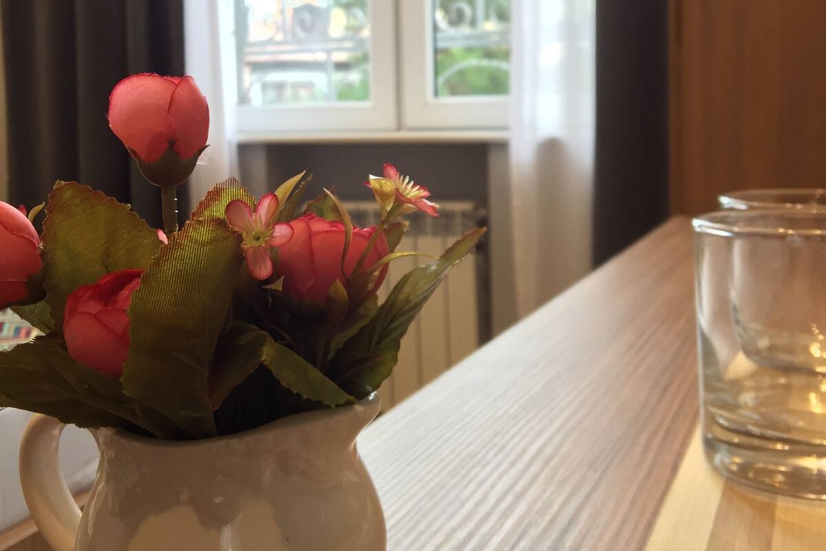 A white ceramic vase with pink flowers on a wooden table near a window with curtains.