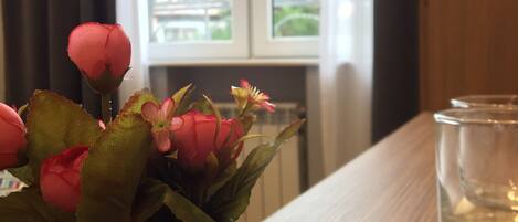 Um vaso de cerâmica branca com flores cor-de-rosa sobre uma mesa de madeira perto de uma janela com cortinas.