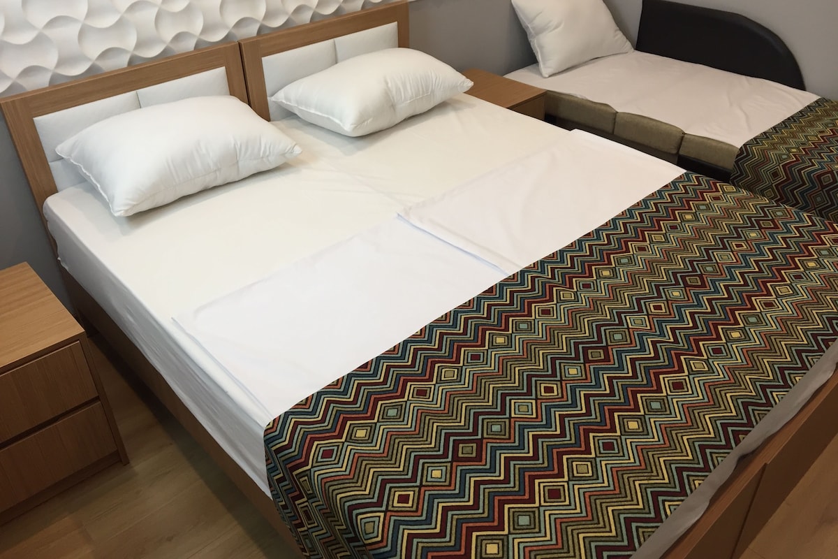 A double bed with white bedding, a patterned throw blanket, and a headboard with a textured design.