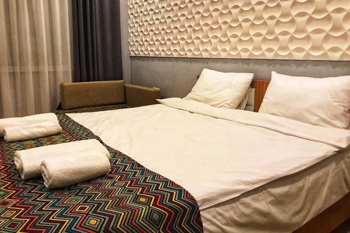 A hotel room with a bed, pillows, a headboard, a sofa, a patterned carpet, and a window with curtains.