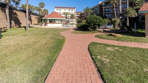 A paved walkway leading to a gazebo in a landscaped area with palm trees and a building in the background. - Seaglass Serenity M112 NEW IN '24 (Galveston)