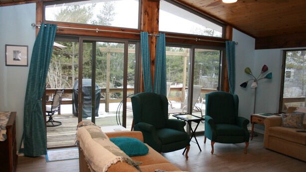 A cozy living room with a wooden ceiling, large windows with curtains, a green armchair, a sofa, and a small table. - Cleves Point Estates Home, Private sandy beach (Greenport)
