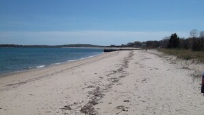 A sandy beach with a wooden pier extending into the water, surrounded by trees and hills in the background. - Cleves Point Estates Home, Private sandy beach (Greenport)