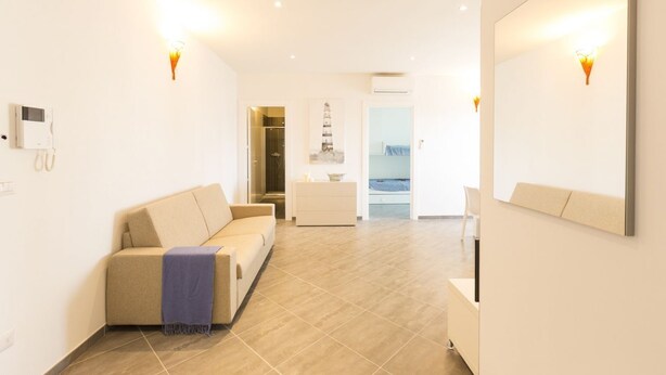 A modern living room with a beige sofa, a small table, and a mirror. There is a door leading to a bathroom and a hallway.