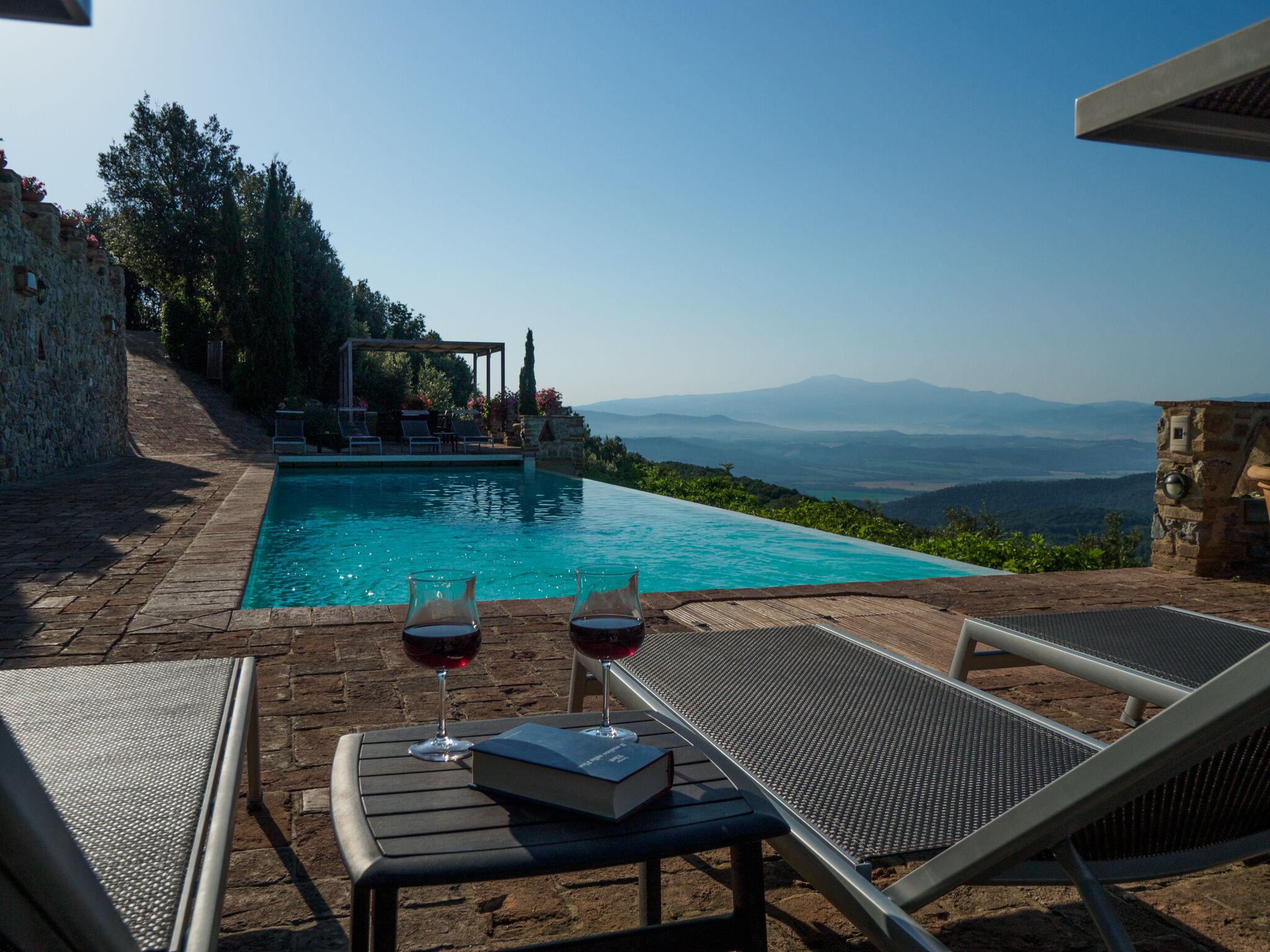 Ein Poolbereich mit Liegestühlen, einem Tisch mit einem Buch und Gläsern Rotwein, mit Blick auf Berge in der Ferne.